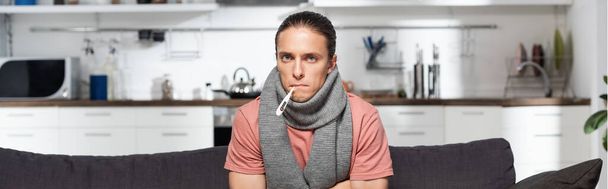 horizontaal beeld van zieke jongeman met warme sjaal op hals en thermometer in de mond kijkend naar camera terwijl hij in de keuken zit - Foto, afbeelding