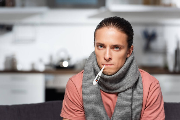 diseased young man with warm scarf on neck and thermometer in mouth looking at camera - Fotoğraf, Görsel