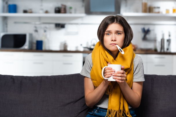 ill woman with thermometer in mouth holding cup of warm tea in kitchen - Foto, immagini