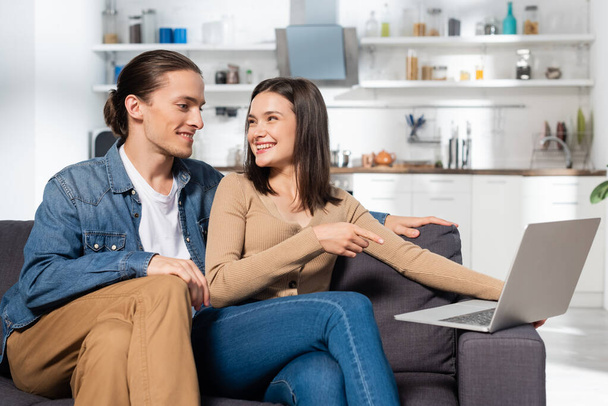 man sitting near excited girlfriend pointing with finger at laptop on couch in kitchen - Φωτογραφία, εικόνα
