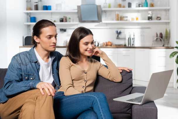 young man and excited woman looking at laptop while sitting on couch in kitchen - Fotoğraf, Görsel