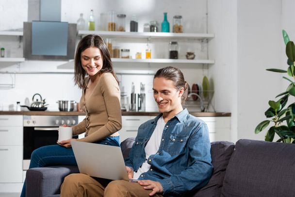 brunette woman with cup of coffee sitting on sofa in kitchen near boyfriend using laptop - Photo, Image