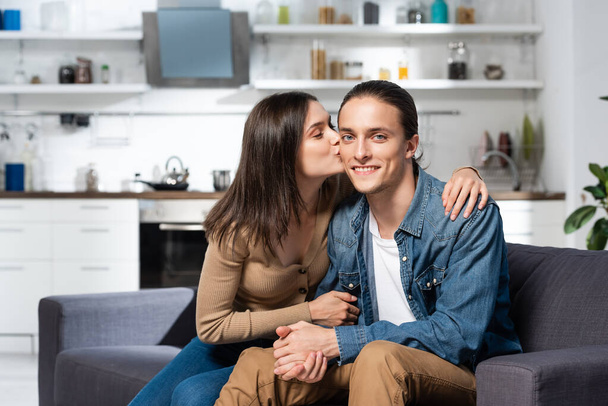 brunette woman kissing joyful man sitting on sofa in kitchen and looking at camera - Photo, image
