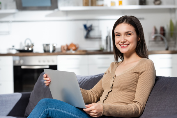 excited freelancer looking at camera while sitting on couch in kitchen with laptop - Photo, Image