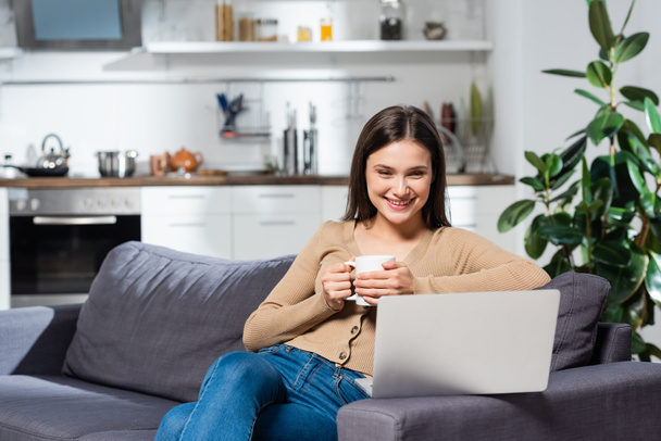 excited freelancer looking at laptop while sitting on sofa in kitchen with cup of warm tea - Foto, Bild