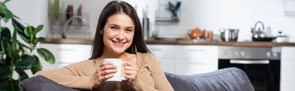 horizontal concept of pleased woman looking at camera while sitting on couch in kitchen with cup of warming beverage - Photo, image