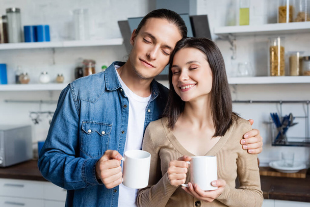 pleased couple standing in kitchen with closed eyes while holding cups of coffee - Fotó, kép