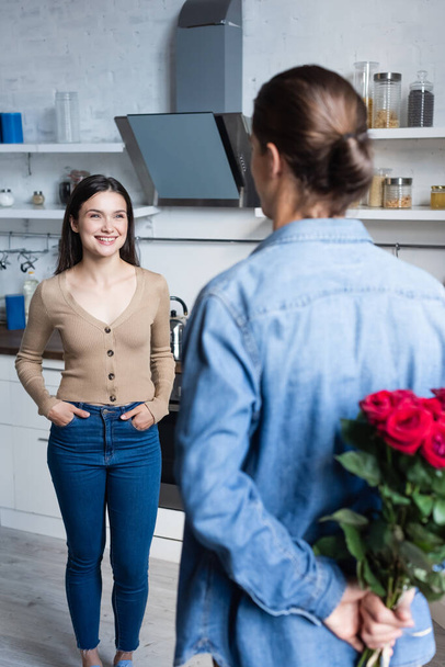 back view of young man holding roses near excited woman standing with hands in pockets in kitchen - Photo, Image