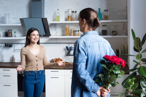 back view of man holding bouquet of roses behind back near excited woman standing with open arms in kitchen - 写真・画像
