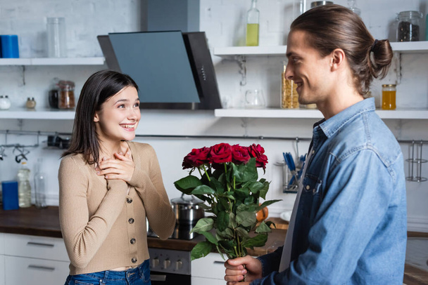 young man presenting bouquet of roses to pleased woman holding hands near chest in kitchen - Photo, Image