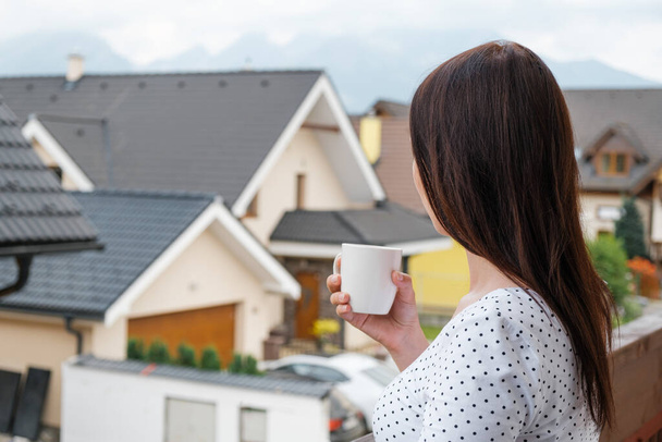Woman standing with a white cup on the balcony and dreamily watching on the cloudy mountain hill. Morning in the countryside. Modern houses near the mountains. advertising concept. Building houses for the key - 写真・画像