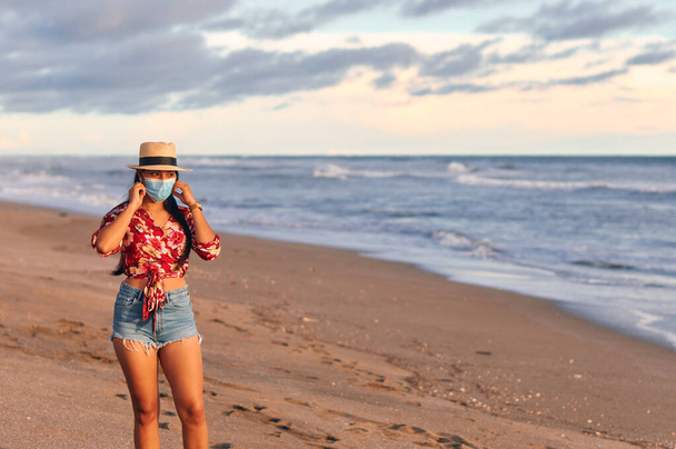 Eine junge Frau trägt ein wunderschönes rotes Hemd mit floralen Mustern, Shorts und Hut und steht mit Gesichtsmaske am Strand, um Covid-19 zu verhindern. Urlaub während der Coronavirus-Pandemie. Neues Normalkonzept - Foto, Bild