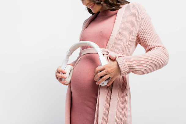 cropped view of pregnant woman holding wireless headphones near belly isolated on white - Zdjęcie, obraz