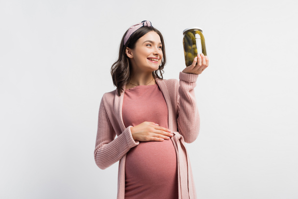 radostná těhotná žena v čelence při pohledu na sklenici s nakládanými okurkami izolovanými na bílém - Fotografie, Obrázek
