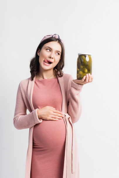 femme enceinte qui sort la langue tout en regardant le pot avec des concombres marinés isolés sur blanc - Photo, image