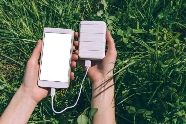 パワーバンクからの充電で少女の手にスマートフォンのモックアップ。緑の畑を背景に - 写真・画像
