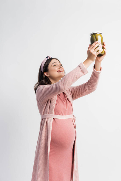 donna soddisfatta e incinta in possesso di vaso con cetrioli sottaceto sopra la testa isolato su bianco - Foto, immagini