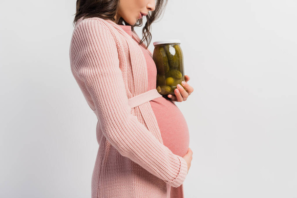 partial view of surprised and pregnant woman holding jar with pickled cucumbers isolated on white - Photo, Image