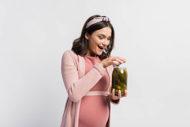 aufgeregte und schwangere Frau öffnet Glas mit sauren und eingelegten Gurken isoliert auf weiß - Foto, Bild