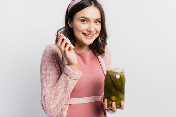 joyful and pregnant woman holding jar with pickled cucumbers isolated on white - Photo, image