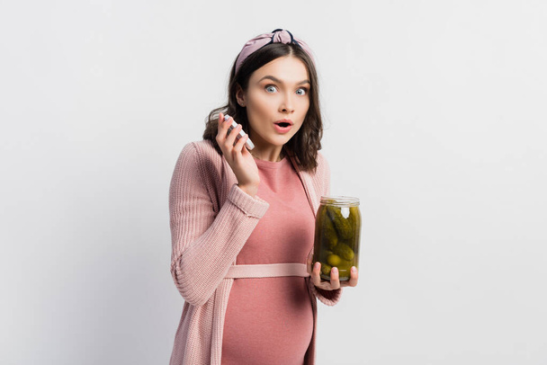 verraste en zwangere vrouw houden pot met gepekelde komkommers op wit - Foto, afbeelding