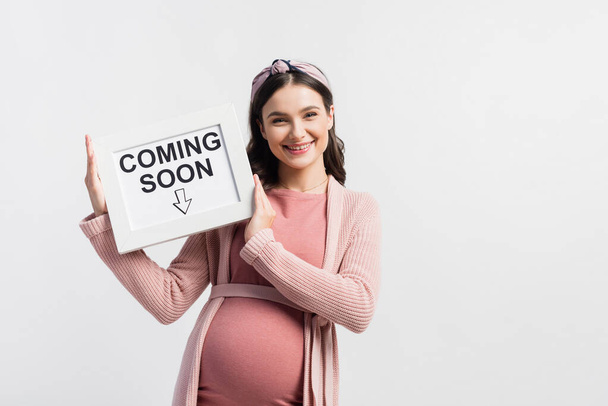 pleased and pregnant woman holding board with coming soon lettering isolated on white - Photo, Image