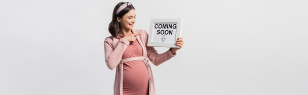 радостная беременная женщина указывая пальцем на доску с приходом скоро буквы изолированы на белом - Фото, изображение