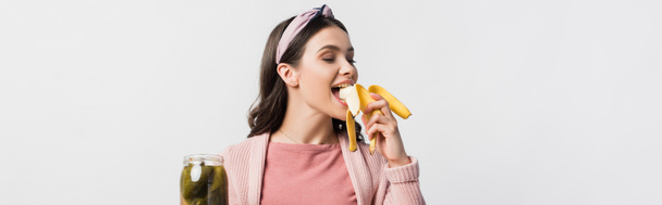 plano panorámico de la mujer comiendo plátano y sosteniendo frasco con pepinos enlatados aislados en blanco - Foto, Imagen