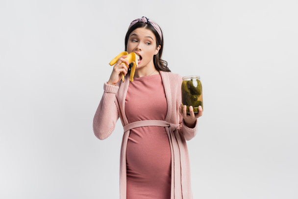 terhes nő eszik banán és nézi üveg konzerv uborka izolált fehér - Fotó, kép
