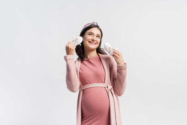 радісна і вагітна жінка тримає крихітні черевики і дивиться вгору ізольовано на білому
 - Фото, зображення