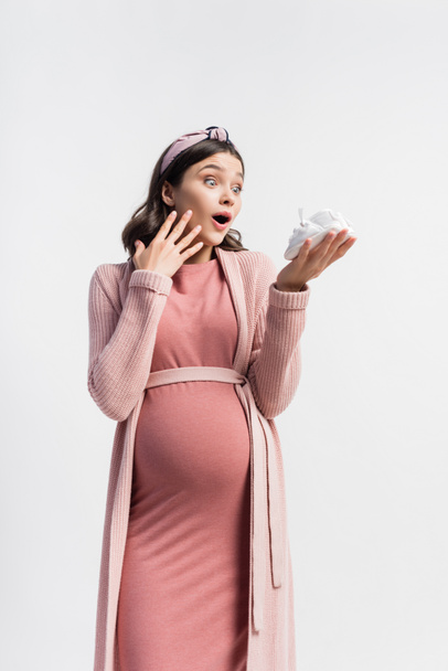 mujer sorprendida y embarazada mirando pequeñas botas aisladas en blanco - Foto, Imagen
