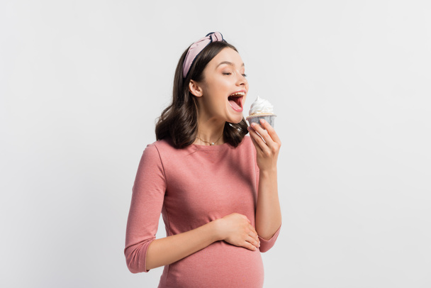 alegre embarazada con boca abierta sosteniendo cupcake aislado en blanco - Foto, imagen