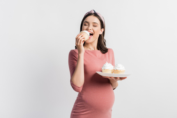 joyful pregnant woman holding plate while eating cupcake isolated on white - Fotoğraf, Görsel