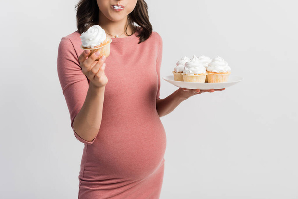 vista recortada de la mujer embarazada sosteniendo plato mientras come cupcake aislado en blanco - Foto, imagen