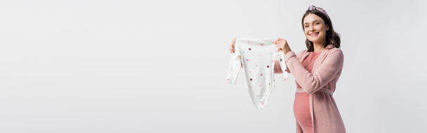 horizontal concept of joyful pregnant woman in headband holding baby romper isolated on white  - Fotó, kép