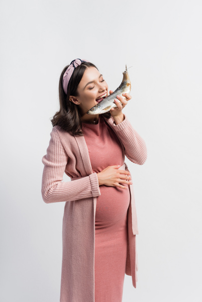 femme enceinte avec les yeux fermés et bouche ouverte manger du poisson séché isolé sur blanc - Photo, image
