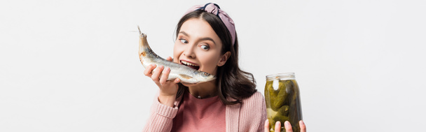 colpo panoramico di donna a bocca aperta con vaso con cetrioli sottaceto e pesce essiccato isolato su bianco - Foto, immagini