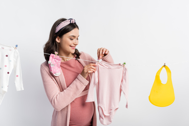 femme enceinte suspendant des vêtements de bébé sur la ligne de vêtements isolés sur blanc - Photo, image