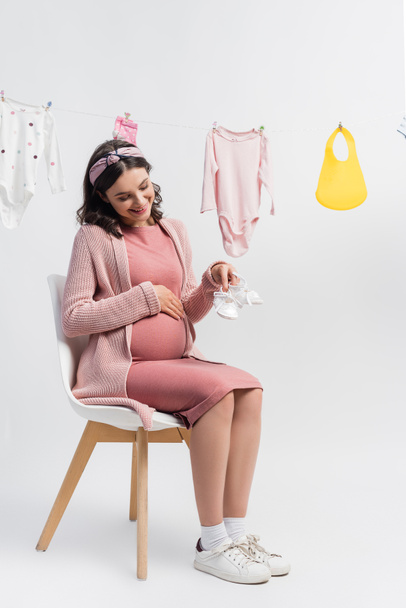 mujer embarazada complacida sosteniendo pequeñas botas y sentada cerca de ropa de bebé en blanco - Foto, Imagen
