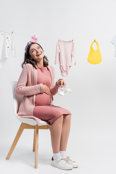 donna incinta e soddisfatta con piccoli stivali e seduta vicino a vestiti per bambini sulla linea di abbigliamento su bianco - Foto, immagini