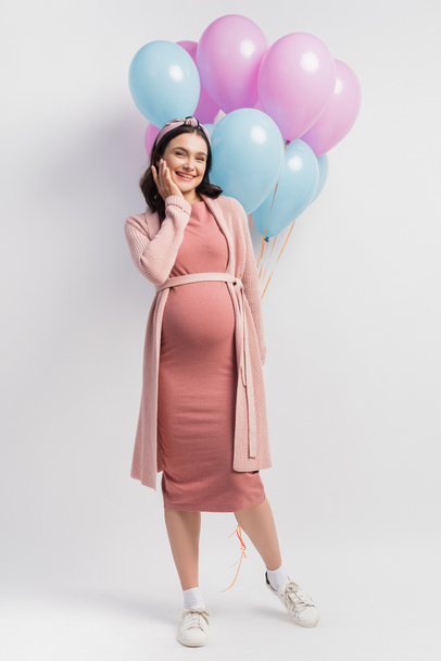 радостная и беременная женщина в платье возле воздушных шаров на белом - Фото, изображение