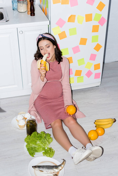 pregnant woman eating banana near jar with canned cucumbers, cupcakes and oranges while sitting on floor  - Foto, Bild