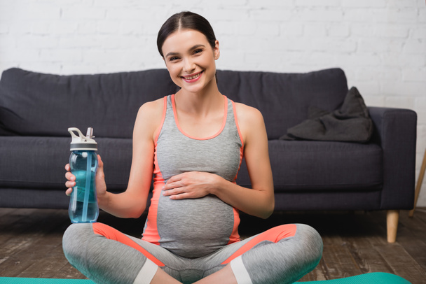 alegre embarazada sentada con botella de deporte en casa - Foto, imagen