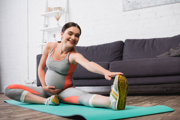 pleased pregnant woman in sportswear stretching on fitness mat - Foto, Bild