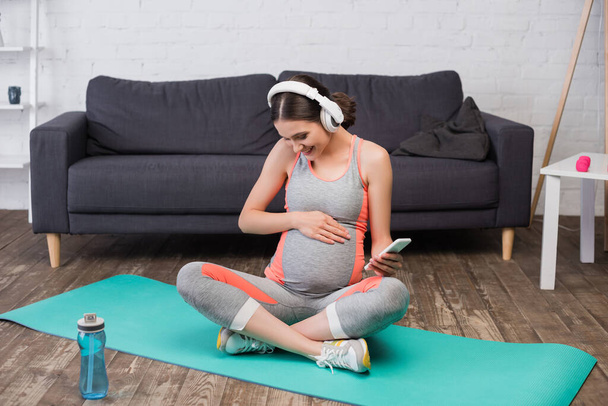 χαρούμενη έγκυος γυναίκα σε ασύρματα ακουστικά ακούγοντας μουσική και κρατώντας smartphone, ενώ κάθεται σε στρώμα γυμναστικής - Φωτογραφία, εικόνα
