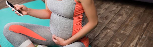 récolte panoramique de femme enceinte tenant smartphone tout en étant assis sur tapis de fitness - Photo, image