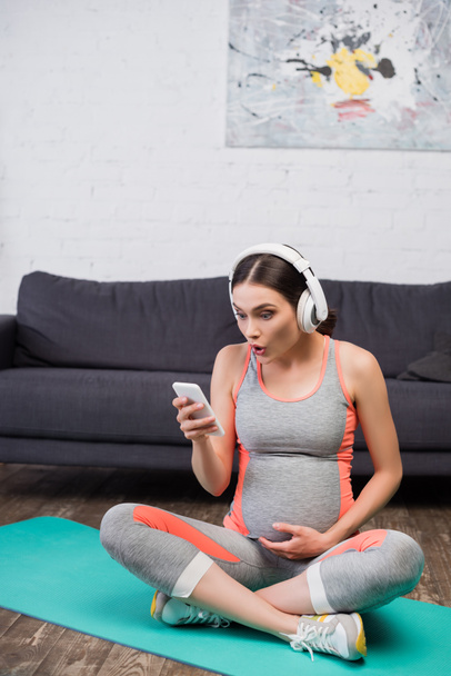 mujer sorprendida y embarazada en auriculares inalámbricos escuchando música y utilizando el teléfono inteligente mientras se sienta en la alfombra de fitness - Foto, Imagen