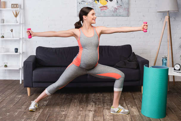 χαρούμενη έγκυος γυναίκα στέκεται με τεντωμένα χέρια, ενώ η άσκηση με ροζ αλτήρες στο σπίτι - Φωτογραφία, εικόνα