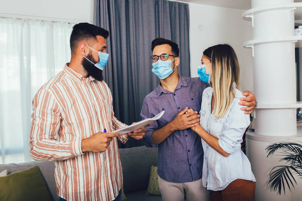 Jeune couple marié heureux dans des masques médicaux debout dans la nouvelle maison avec agent immobilier masculin. Concept pandémique. - Photo, image