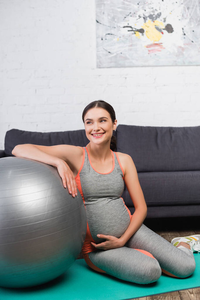 donna gioiosa e incinta toccare la pancia mentre seduto vicino palla fitness in soggiorno  - Foto, immagini